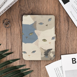 May include: A rectangular e-reader case featuring a repeating pattern of cat faces in various colors including blue, cream, white, and grey. The cats have distinct eye colors such as blue, green, and yellow. The case has a smooth, matte finish.