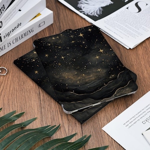 May include: Black and gold tablet case with a celestial design. The case features a dark background with gold stars and a wavy, layered pattern. The case is lying on a wooden surface.