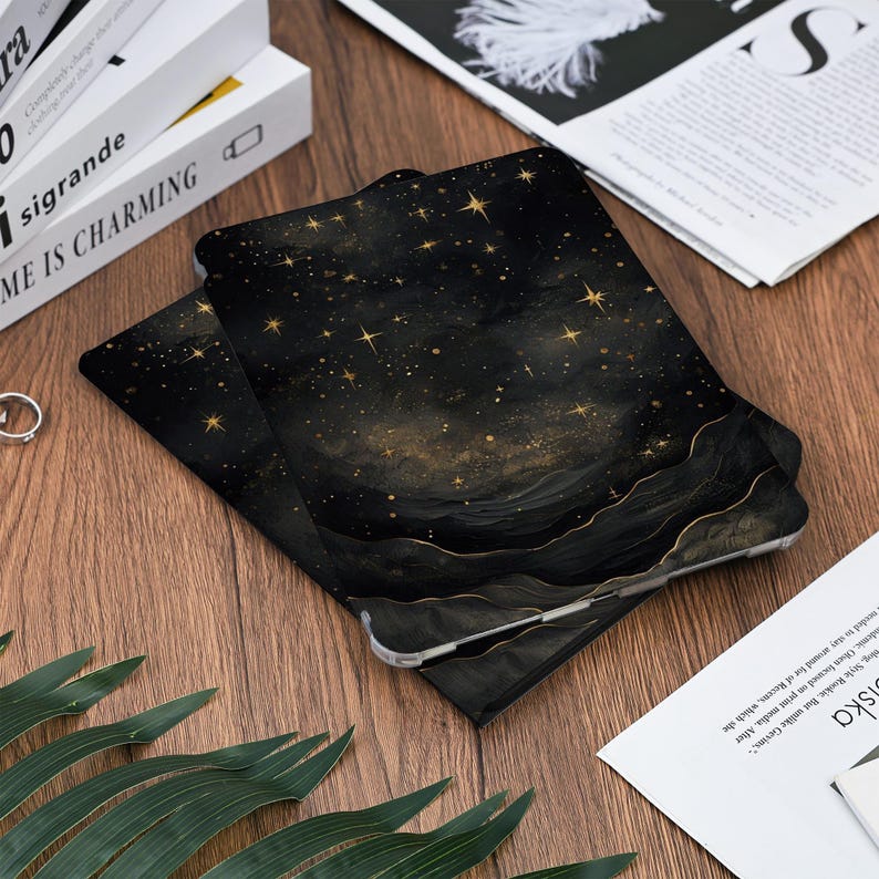 May include: Black and gold tablet case with a celestial design. The case features a dark background with gold stars and a wavy, layered pattern. The case is lying on a wooden surface.
