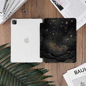 May include: A clear iPad with a protective case next to a black and gold galaxy-themed iPad case. The galaxy case features a starry night design with gold stars and a dark, swirling background. The iPad is on a wooden surface.