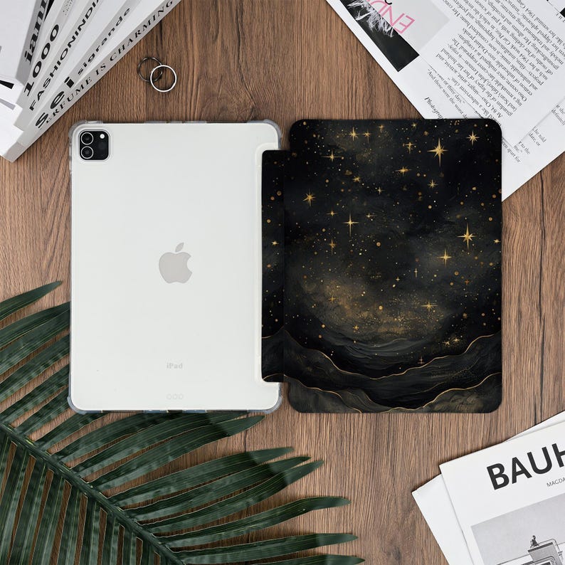 May include: A clear iPad with a protective case next to a black and gold galaxy-themed iPad case. The galaxy case features a starry night design with gold stars and a dark, swirling background. The iPad is on a wooden surface.