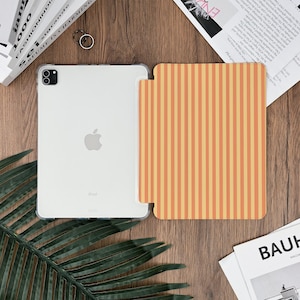 Pu&ograve; includere: Una custodia trasparente per iPad e una cover a righe verticali arancioni e gialle sono esposte su una superficie di legno. L'iPad ha un logo Apple argentato. Sono visibili anche un anello e una rivista.