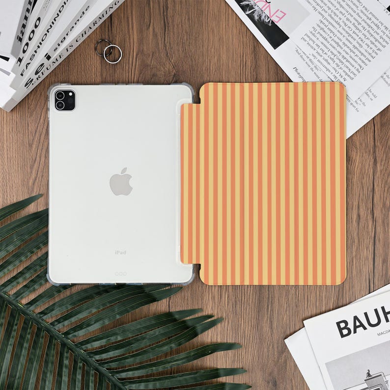 Pu&ograve; includere: Una custodia trasparente per iPad e una cover a righe verticali arancioni e gialle sono esposte su una superficie di legno. L'iPad ha un logo Apple argentato. Sono visibili anche un anello e una rivista.
