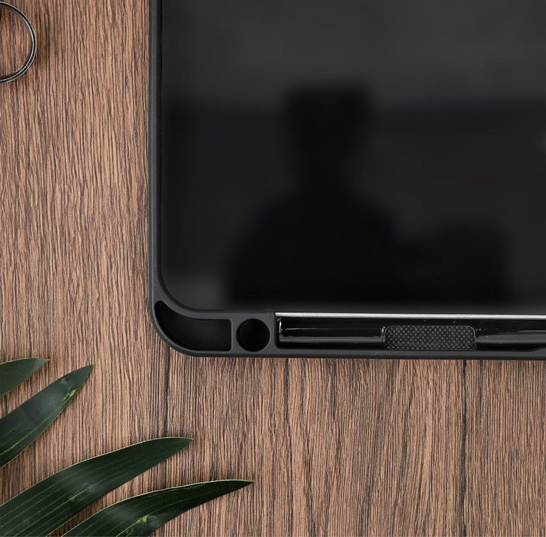 Pu&ograve; includere: Una custodia per telefono nera con finitura opaca. La custodia ha un design elegante con angoli arrotondati e un pulsante strutturato. La custodia &egrave; su una superficie di legno con foglie verdi.