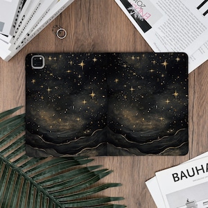 May include: Black and gold tablet case featuring a starry night design. The case has a dark background with gold stars and a wavy, mountain-like pattern at the bottom. The case is lying on a wooden surface.