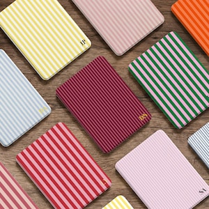 Puede incluir: Una colección de cuadernos rectangulares con rayas verticales en varios colores, incluyendo amarillo, rosa, rojo, verde y azul. Cada cuaderno tiene un monograma dorado en la esquina. Los cuadernos están dispuestos sobre una superficie de madera.