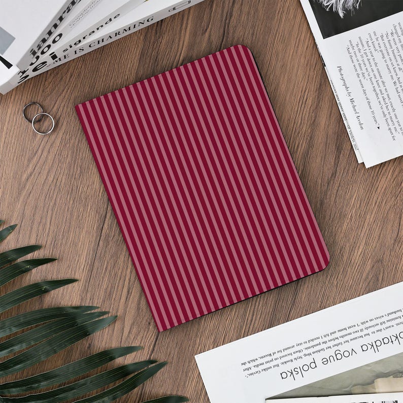May include: A rectangular notebook with a vertical striped pattern in shades of red and pink. The notebook is lying on a wooden surface, with a few books and a magazine visible in the background.