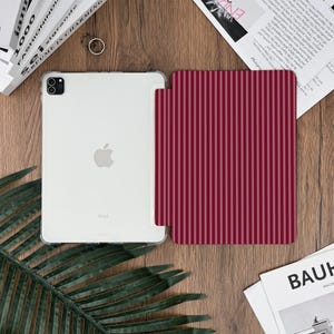 May include: A clear iPad with a protective case and a burgundy striped cover. The iPad is white with the Apple logo visible. The cover has vertical stripes in varying shades of burgundy. The items are on a wooden surface.