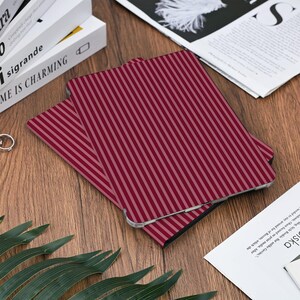 May include: A burgundy and light pink striped tablet case is displayed on a wooden surface. The case is rectangular with rounded corners and a vertical stripe pattern. Other items on the surface include books, a newspaper, and greenery.