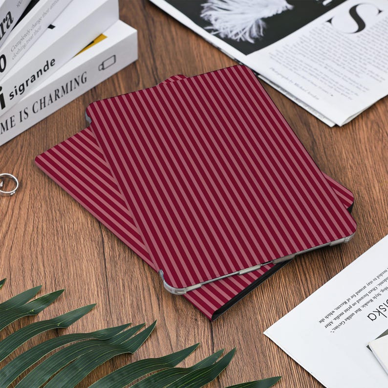 May include: A burgundy and light pink striped tablet case is displayed on a wooden surface. The case is rectangular with rounded corners and a vertical stripe pattern. Other items on the surface include books, a newspaper, and greenery.