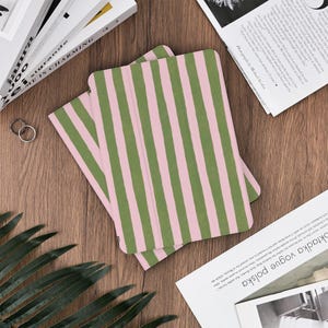 Pu&ograve; includere: Due custodie per tablet con un motivo a righe rosa e verdi. Le custodie sono aperte, rivelando l'interno. Sono su una superficie di legno, con libri e riviste sullo sfondo.