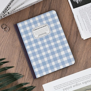 Peut inclure: Un cahier de composition à carreaux bleus et blancs. Le cahier porte une étiquette blanche avec l'inscription "COMPOSITION BOOK" et des détails sur le nombre de feuilles et les dimensions. Le cahier est posé sur une surface en bois.