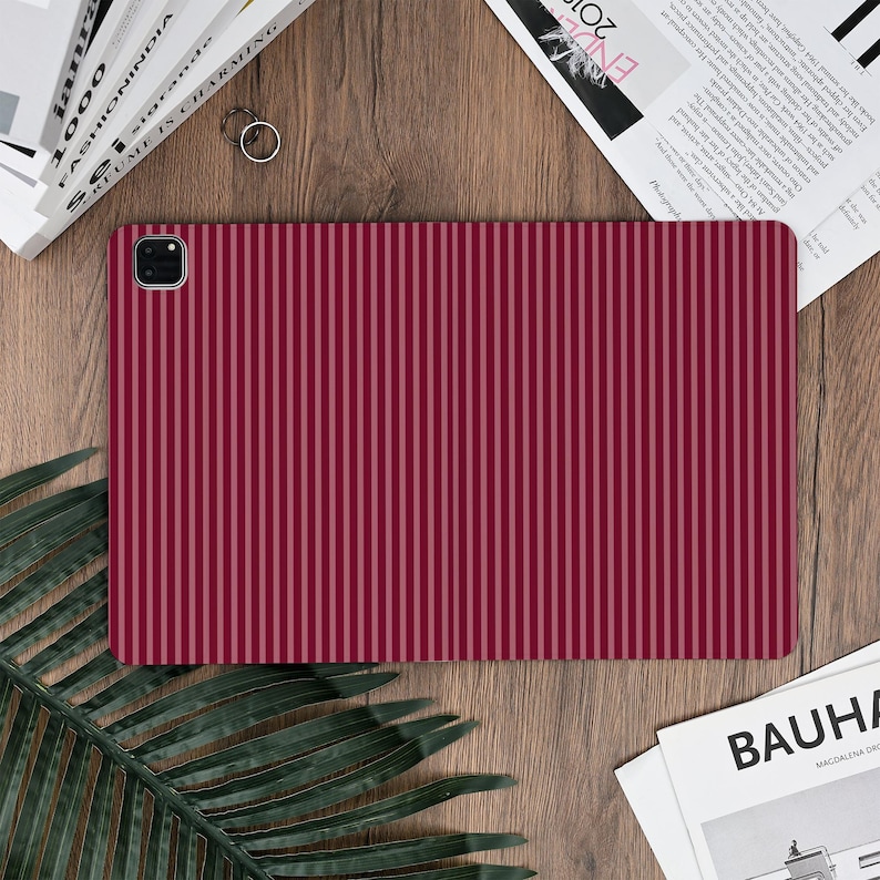 Pu&ograve; includere: Una custodia per tablet bordeaux con strisce verticali in varie tonalit&agrave; di rosso. La custodia ha un ritaglio per la fotocamera ed &egrave; appoggiata su una superficie di legno con libri e una rivista sullo sfondo.