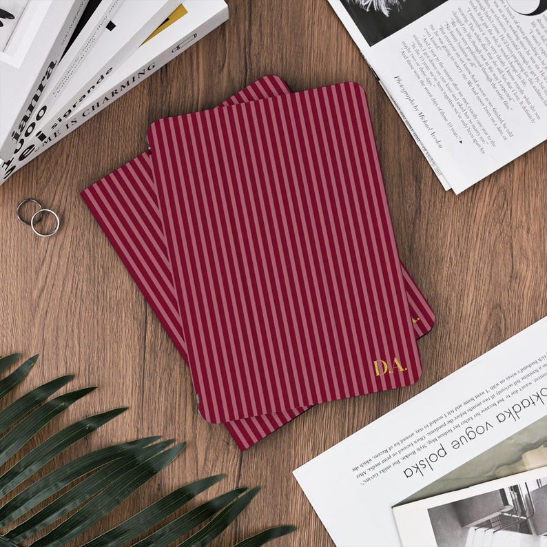 May include: Two burgundy and pink striped notebooks with gold initials "D.A." are displayed on a wooden surface. The notebooks are open, revealing the striped pattern. Other items include books, magazines, and rings.