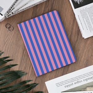 Pu&ograve; includere: Un taccuino con un motivo a righe rosa e blu. &Egrave; appoggiato su una superficie di legno, con altri oggetti come libri e anelli nelle vicinanze. Il testo "ME IS CHARMING" &egrave; visibile su un libro.