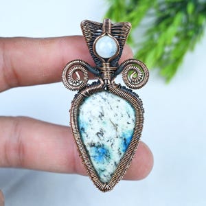 Può includere: Un ciondolo a forma di goccia con una pietra screziata bianca e blu, avvolta in un intricato filo di rame ossidato. Il design presenta due spirali laterali simmetriche e un piccolo cabochon rotondo di colore bianco traslucido in alto.