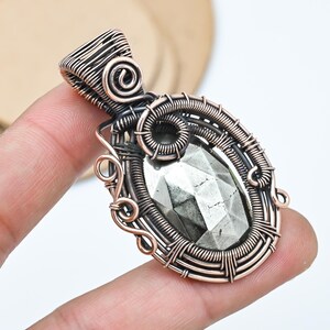 May include: A handcrafted pendant featuring a faceted, oval-shaped pyrite stone, intricately wrapped in copper wire. The wirework includes decorative swirls and a bail for a necklace chain. The pyrite has a metallic, silver-gray color.