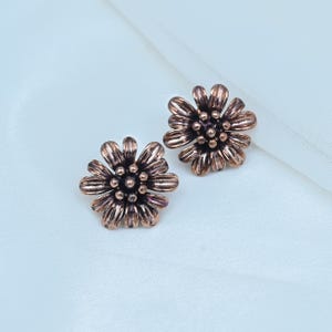 May include: A pair of copper-toned metal stud earrings shaped like daisies. Each earring features a central cluster of small spheres surrounded by multiple elongated, textured petals radiating outward in a circular floral pattern.