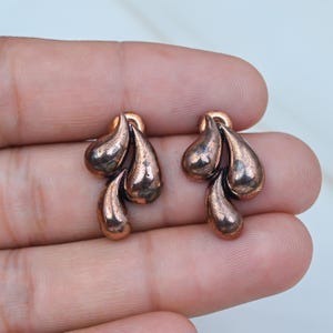 May include: A pair of copper-toned metal earrings, each featuring a cluster of three smooth, teardrop-shaped elements arranged in a cascading formation with a polished, reflective surface finish.