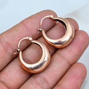 Puede incluir: Un par de pendientes de aro pequeños y gruesos fabricados con un material metálico en tono cobre. Los pendientes presentan un perfil liso, redondeado y en forma de gota con un mecanismo de poste articulado para un cierre seguro y acabado.