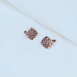 May include: A pair of square-shaped copper-toned earrings featuring a textured, cross-hatched grid pattern on the front surface. Each earring is equipped with a hinged lever-back closure mechanism visible on the side of the square metal piece.
