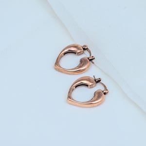 May include: A pair of copper-toned hoop earrings shaped like hearts. Each earring features a smooth, polished metallic finish with a hinged clasp mechanism at the top, creating a continuous, rounded heart silhouette for the jewelry piece.