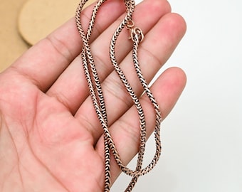 May include: A copper-colored chain necklace with a woven design. The necklace is held in a hand, showcasing the intricate pattern and the warm metallic hue. The clasp is visible, indicating the necklace is ready to wear.