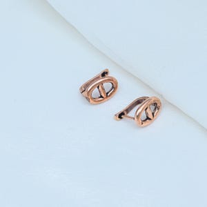 May include: A pair of copper-toned earrings featuring an oval-shaped frame with a central horizontal bar. The earrings have a hinged lever-back closure mechanism and a textured, slightly darkened finish on the metal surface.