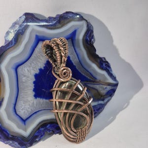 May include: A handcrafted pendant featuring a polished, oval-shaped stone wrapped in copper wire. The stone has a dark green hue with subtle variations. The copper wire is intricately woven around the stone, creating a cage-like design. The pendant is displayed on a blue and white agate slice.