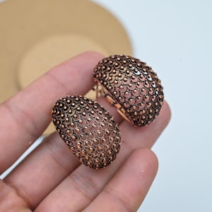 May include: Two copper-toned, oval earrings. Each earring features an intricate openwork design with tiny raised dots and circular patterns. The earrings are presented against a light backdrop, highlighting their detailed texture and warm metallic colour.