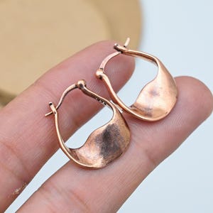 May include: A pair of copper-coloured hoop earrings. The earrings have a unique, curved design with a flattened, textured surface. The earrings are displayed on a light background.