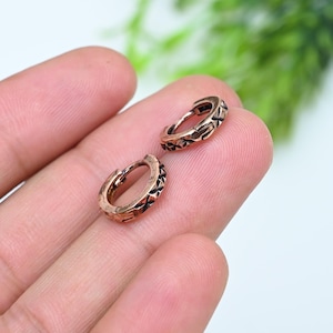 May include: A pair of small, circular hoop earrings crafted from a copper-toned metal. Each earring features a repeating engraved pattern of small, dark-filled crosses or stars along the outer surface, with a hinged closure mechanism.