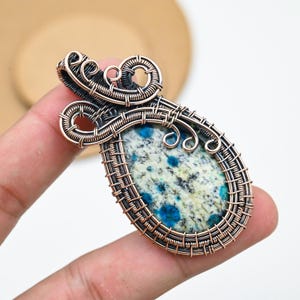 Può includere: Ciondolo fatto a mano con una pietra a forma di goccia con motivi blu e bianchi, incastonata in un'intricata filatura di rame. La filigrana include volute decorative e un bordo intrecciato, creando un gioiello unico e artistico.
