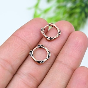 May include: A pair of small, circular hoop earrings crafted from a rose gold-toned metal. Each hoop features a textured, cross-hatched pattern along the outer surface, creating a subtle, repeating geometric design across the metallic finish.