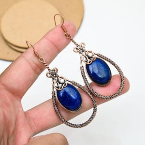 以下が含まれることがあります： 手作りの銅製イヤリング。涙の形をしたフレームが特徴で、各イヤリングの中央には濃い青色の楕円形の石があります。銅線は石とフレームの周りに複雑に織り込まれ、ユニークで芸術的なデザインを作り出しています。