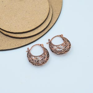 May include: A pair of copper-coloured hoop earrings with an open filigree design. The earrings are crescent-shaped and feature a detailed, interwoven pattern. They are set against a white background with a light brown surface in the upper left corner.