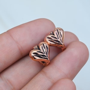 May include: Two small, heart-shaped copper beads with a metallic finish. Each bead features a series of diagonal, incised lines carved into the surface, creating a textured, fan-like pattern across the rounded heart shape.