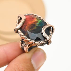 May include: A copper-toned wire-wrapped ring featuring a large, faceted gemstone with a vibrant display of colors, including red, orange, green, and blue. The ring's intricate wirework frames the stone, creating a unique and artistic piece of jewelry.