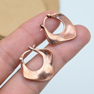 May include: A pair of copper-colored square hoop earrings. The earrings have a slightly textured surface and a warm, reddish-brown hue. The design is a modern take on a classic hoop, with a squared-off shape and a simple, elegant style.