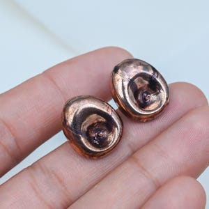 May include: A pair of stud earrings with a metallic copper finish. Each earring features an irregular, rounded, organic shape with a smooth, reflective surface and a central indentation where the post is attached to the back of the piece.