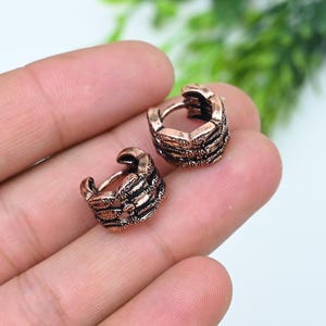 May include: A pair of small hoop earrings with a copper-toned finish and a textured, segmented design resembling a skeletal structure. The earrings feature a hinged mechanism and a dark, oxidized patina within the recessed areas of the metal.