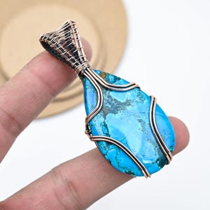 以下が含まれることがあります： 鮮やかなターコイズストーンを銅線で精巧に包んだ、涙型のペンダント。青い石には濃い色の縞模様があり、銅線は上部で織り込まれて安全なベイルになっています。ユニークなジュエリーです。