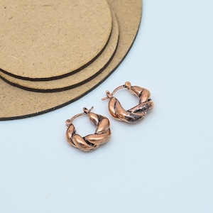 May include: Two copper-toned hoop earrings featuring a woven design. The earrings are small and have a textured finish. The background is a light blue surface with some brown circular objects.