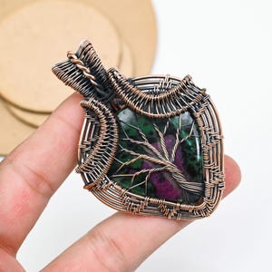 Puede incluir: Un colgante con una piedra de zoisita pulida en forma de escudo con inclusiones verdes y magenta. La piedra está envuelta en un intrincado alambre de cobre oxidado que forma un diseño de árbol sobre la superficie y una argolla.
