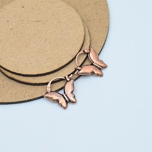 Puede incluir: Dos pendientes con forma de mariposa de color cobre. Cada pendiente presenta un diseño de mariposa con patrones de alas detallados. Los pendientes se muestran sobre una superficie circular marrón claro.