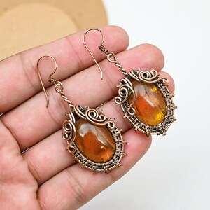 May include: Handmade copper wire earrings with oval amber-coloured stones. The stones are set in intricate wirework with decorative swirls and a woven border. The earrings have a hook closure and a twisted wire design.