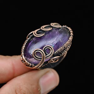 Puede incluir: Un anillo ovalado de amatista con alambre de cobre. El anillo presenta una piedra púrpura grande y pulida engastada en un diseño de alambre detallado y hecho a mano. El trabajo de alambre incluye remolinos decorativos y una banda texturizada.