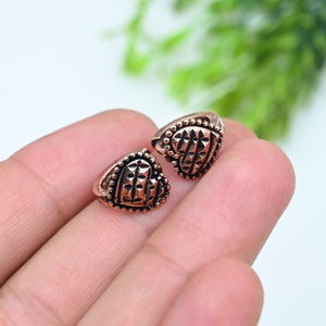 May include: Two small copper-toned heart-shaped charms featuring a textured surface with a series of vertical indentations and a beaded border. The charms have a metallic finish and a distinct, raised pattern across the center of each heart shape.