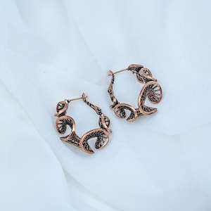 Puede incluir: Un par de pendientes de aro hechos de metal en tono cobre, con un intrincado diseño de filigrana calada con patrones de volutas. Los pendientes cuentan con un cierre de poste articulado y un acabado oscuro y envejecido en las zonas hundidas.
