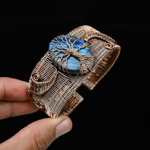 Puede incluir: Brazalete de alambre de cobre con una piedra preciosa azul central con un diseño de árbol de la vida. El brazalete tiene un diseño abierto y detalles intrincados de alambre tejido. La piedra preciosa mide unos 2,5 cm de diámetro.
